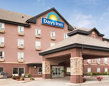 A small photo of the parking lot entrance to the d3h Days Inn Calgary Airport hotel, a four storey light terracotta building with the Days Inn yellow sunrise and blue background corporate logo placed at the triangular summit of the hotel. A large concrete portico with stone accents and triangular shaped roof provides coverage over the glass door entrance.