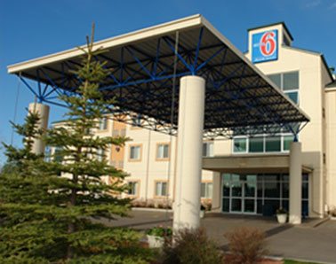 A small view of the glass door entrance to d3h Motel 6 in Red Deer, with two green balsam fir trees placed at the foot of a rectangular portico supported by concrete posts. The hotel's corporate logo (represented by the number 6 in red bold type contrasted against a blue background) is placed at the summit of the three storey building above the entrance.