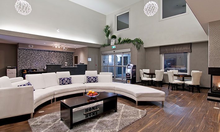 The hotel lobby of d3h Home Inn & Suites Regina Airport features hardwood flooring and a semi-circular white sectional sofa, encircling a solid dark wood coffee table placed over an area rug.  The charcoal gray check-in counter is contrasted against a white and dark patterned accent back wall, while the ATM and two square tables, complemented by white upholstered Parsons chairs, are set at the periphery of the lobby.