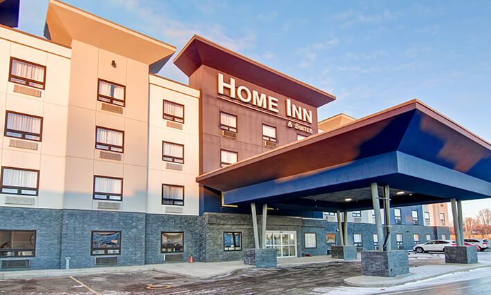 The exterior parking lot view of the four storey building of d3h Home Inn & Suites Saskatoon South on a snowy day in Saskatchewan.  A large concrete portico with four steel and concrete posts provide coverage over the back entrance to the hotel, while several parked cars can be seen in the distance.