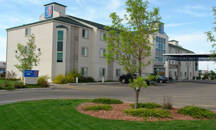 A side view of the parking lot and back entrance to the d3h Motel 6 in Red Deer, Alberta.  In the foreground, a landscaped area features a tree surrounded by manicured lawn space.  Two vehicles are parked next to the concrete and steel portico covering the entrance to the motel.  The corporate logo (in red, white and blue), used as signage, is placed in two locations at the summit of the ivory beige three storey building.