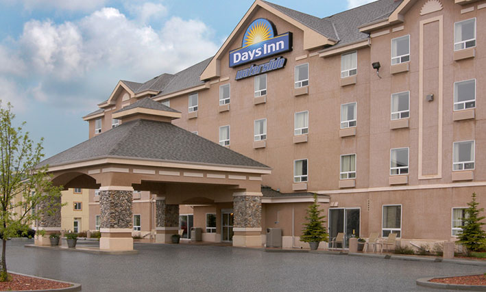 The entrance view of d3h Days Inn Red Deer hotel in Alberta showcases a large square pavilion-shaped portico constructed of concrete and stone.  Placed at the summit of the four storey hotel is the Days Inn corporate sign (represented with a yellow sunrise icon against a blue background), accompanied by lettering signage with waterslide presented in blue and white outline typeset.