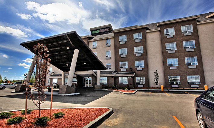 The d3h HomeSuites Regina East hotel is a four storey building painted in two tone chocolate brown and beige.  The HomeSuites corporate name is presented in red and green lettering signage positioned at the summit of the hotel.  The parking lot is relatively empty with two vehicles parked nearby the tilted rectangular portico supported with concrete columns.