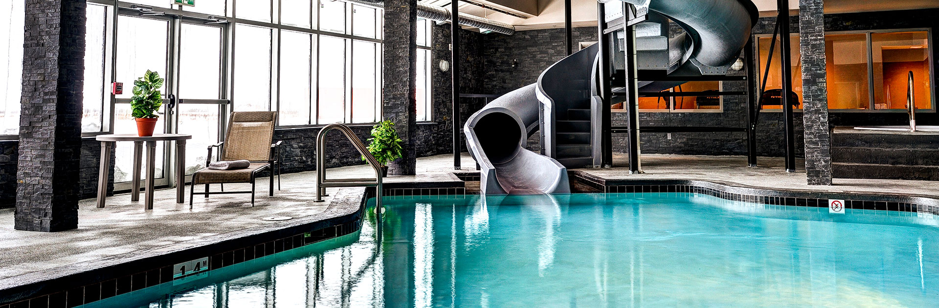 The pool and waterslide area at d3h Home Inn & Suites in Swift Current is bathed in natural light from large picture metal framed windows, shining onto charcoal gray stone walls and posts. A small lounge area at the edge of the pool is furnished with a wicker patio chair and a sidetable on which a plant is placed.