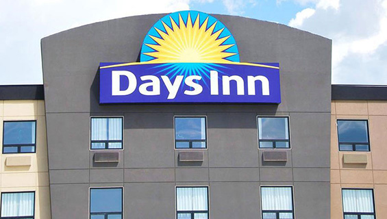 A large scale sign of the Days Inn logo is placed high atop the roof of the graphite-gray exterior of a Days Inn property.  The Days Inn corporate name is presented in bold white type, with a yellow sunrise icon contrasted against a blue background.