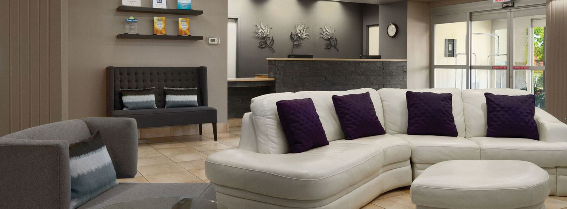 The lobby in d3h Days Inn Edmonton South is a welcoming space with comfortable seating:  a plush white sectional sofa with matching ottoman, a gray single seat armchair and charcoal gray loveseat placed next to the stone check-in counter.  Three silver and black metallic tulip wall ornaments are mounted against the cloudy concrete gray walls behind the check-in desk.