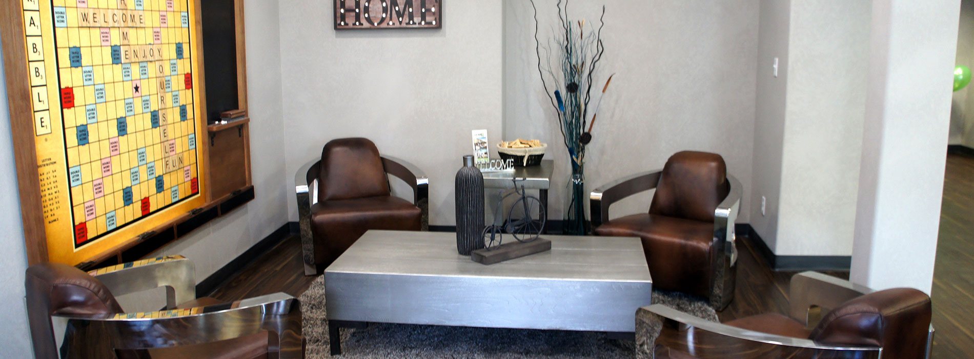 Four leather club chairs with thick metallic arms are matched with a solid metal top coffee table and side end table in the lounge and recreation space of d3h Home Inn Express in Medicine Hat, Alberta.  A large Scrabble board game is mounted against light gray walls with an attached blackboard for score keeping.