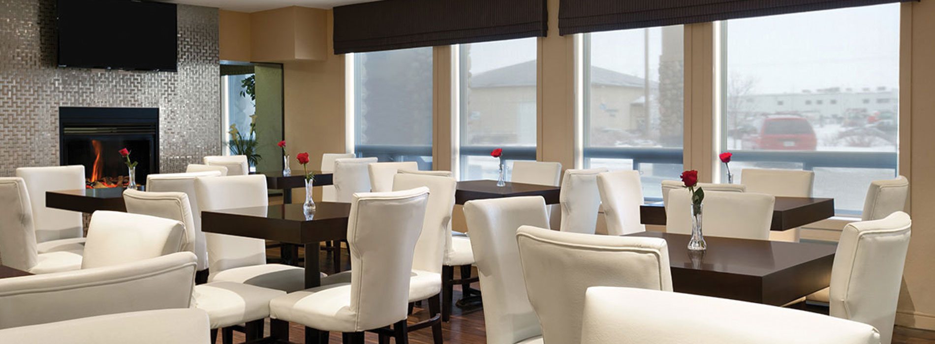 Natural light from large windows in the dining area at d3h Days Inn Regina, reflect off the shiny metallic surround of the glass enclosed fireplace, with a flatscreen TV mounted above.  Single red roses in bud vases sit atop square eating tables surrounded by white upholstered dining chairs.