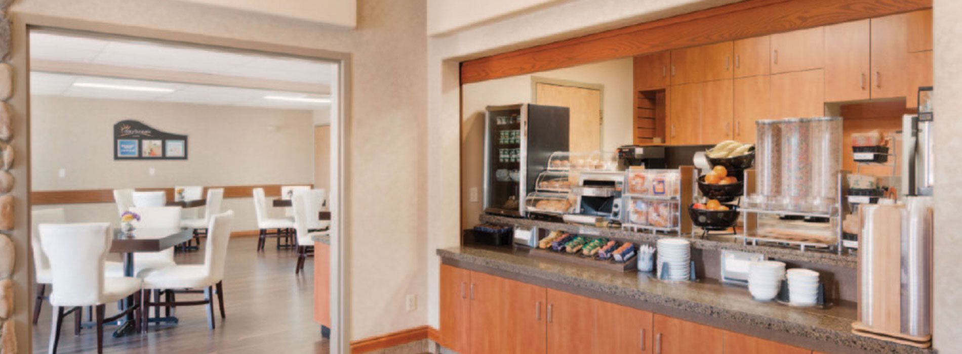 The Daybreak Café at Days Inn Red Deer showcases a long granite top counter with red ochre brown cabinetry.  Bowls of fruit, a mini refrigerator unit, stainless steel toaster, baked goods housed in display cases, and dining ware all sit atop the breakfast bar, which overlooks the entrance to the eating space populated by square tables and white upholstered Parsons chairs.