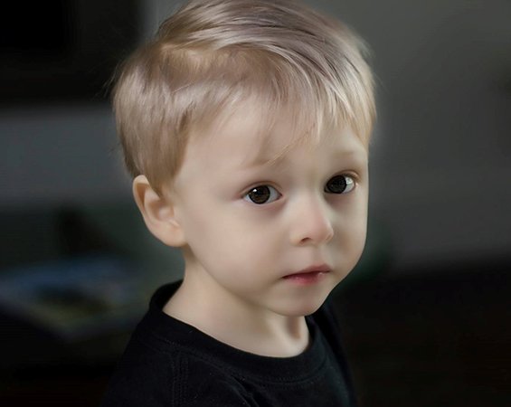 A smiling blond male toddler dressed in a seafoam green jumper, looks out of a large glass window on a bright and sunny day.