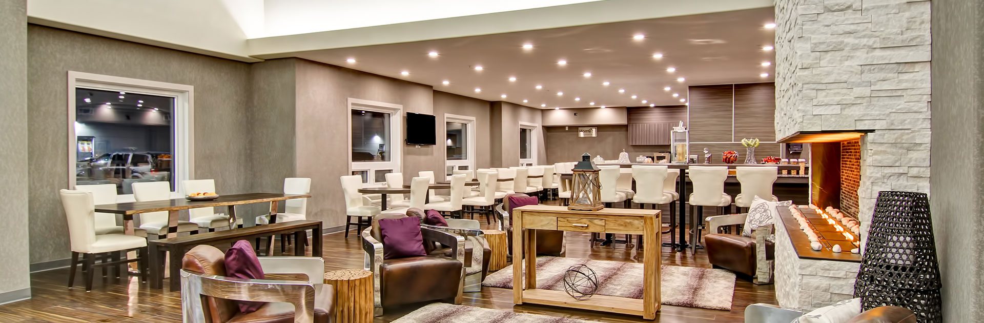 An elegantly designed eating and lounging area at a d3h managed hotel property showcases dark wood eating and dining tables encircled by white upholstered chairs and a large white stone fireplace accompanied by four leather lounge chairs.