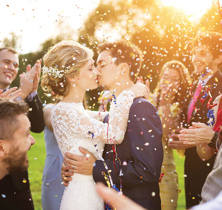A newlywed couple (the bride dressed in white lace, the groom in a blue suit) kiss as friends and family happily applaud while confetti rains down all around.