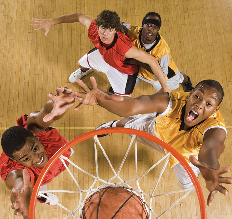 Four young males engaged in a game of basketball, stop and watch in anticipation as the ball is shot into the basket and the point is scored.  