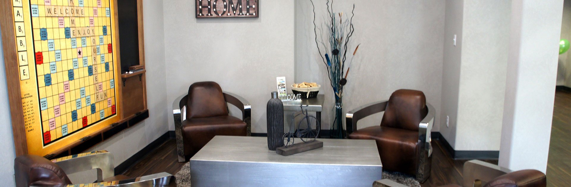 The recreation space at d3h Home Inn Express in Medicine Hat, Alberta, showcases a wall mounted large Scrabble game board with attached blackboard overlooking four leather club chairs placed amongst matching steel topped coffee and end tables.  A small HOME sign hangs on the adjacent wall.