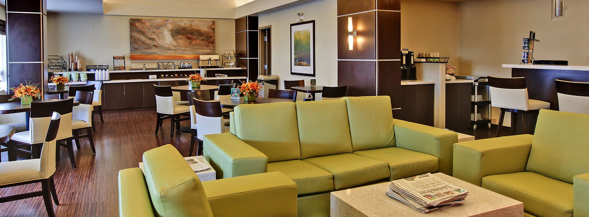 The breakfast and sitting space at d3h Home Inn & Suites Yorkton features wood grain chestnut brown hardwood floors and a long two tiered breakfast counter stocked with dining ware and stainless steel appliances.  Square eating tables and upholstered dining chairs are placed throughout the breakfasting area.  In the sitting area, modern cube shaped coffee and end tables flank a chartreuse green vinyl sofa and armchair set.