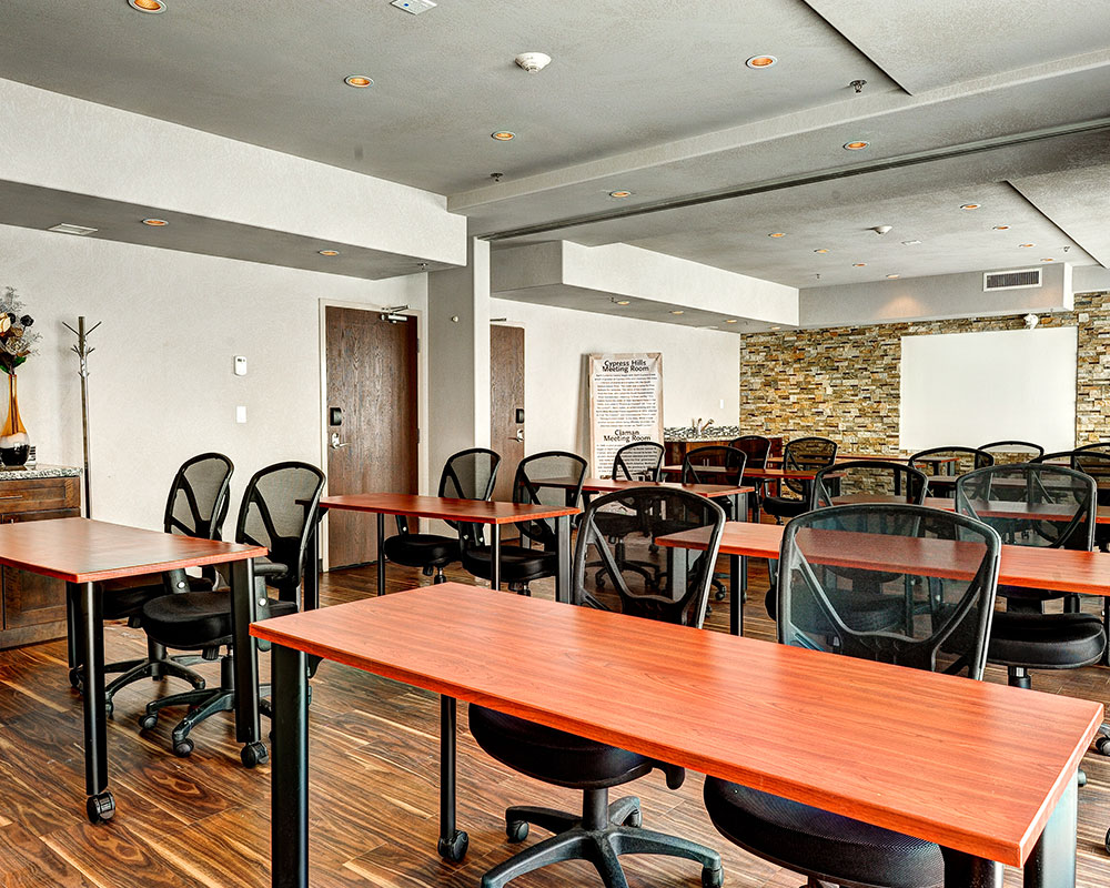 Rows of red cherry wood writing desks, each with a pair of black ergonomic chairs, are placed throughout the hardwood floor space of the meeting room at d3h Home Inn & Suites Regina Airport.  A white board is mounted against a vivid multi-color stacked stone accent wall at the back of the room.