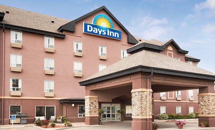 A large square pavilion shaped portico constructed of concrete with stone accents, covers over the glass door entrance to d3h Days Inn Calgary Airport hotel in Alberta.  The Days Inn corporate logo (represented by a yellow sun with a blue background) is placed at the triangular roof summit of the four storey building.  To the side of the entrance, sits a barbecue grill and an outdoor lounge area furnished with patio chairs.