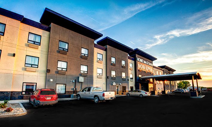 A small view of the back parking lot of d3h Home Inn & Suites Yorkton with several vehicles parked up to the three storey, two tone slate-gray and off-white building.  A semi-circle shaped portico hovers over the hotel entrance with the corporate name in presented in white lettered signage placed above.  A large orange banner inscribed in bold white lettering Now Open is draped beneath the corporate sign.