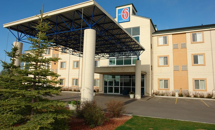 The parking lot entrance to d3h Motel 6 Red Deer features a concrete and steel portico with support posts providing coverage over the glass entrance to the three storey hotel.  Attached to the rooftop summit of the two-tone pastel yellow and latte building, the squared shaped Motel 6 logo (with red and white typeset against a blue background) is used as signage.