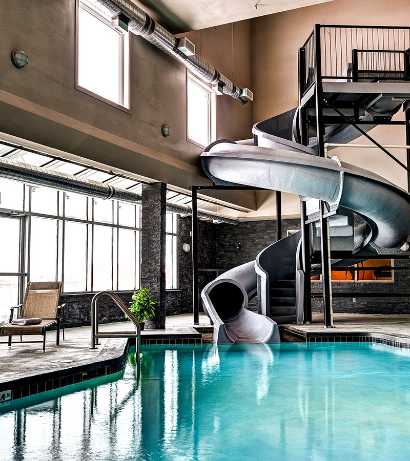 Abundant natural lighting flow in from large windows and glass doors of the indoor swimming area at d3h Home Inn & Suites Swift Current, revealing a twisting large gray waterslide meandering into the black tiled pool.  A lone wicker chaise lounge is placed by the pool's edge for spectators.