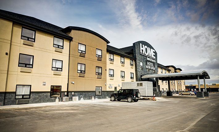 The view of the parking lot of the d3h Home Inn & Suites Swift Current hotel on a clear day, with several vehicles parked next to the semi-circular metal portico covering over the entrance.  The corporate name (in large white lettering) is placed above the entrance, with a black banner advertising Hotel Now Open in bold white lettering typeset.