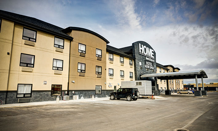 Several cars are parked near the back entrance to the d3h Home Inn & Suites Swift Current hotel, which is covered by a semi-circular portico supported by steel posts encased in a stone and concrete base.  The three storey hotel building is painted in tri-tone slate gray, cream and tan.  Above the hotel entrance, Home Inn & Suites is displayed as signage in large and prominent white lettering with an accompanying black banner beneath, announcing Hotel Now Open in bold white typset.