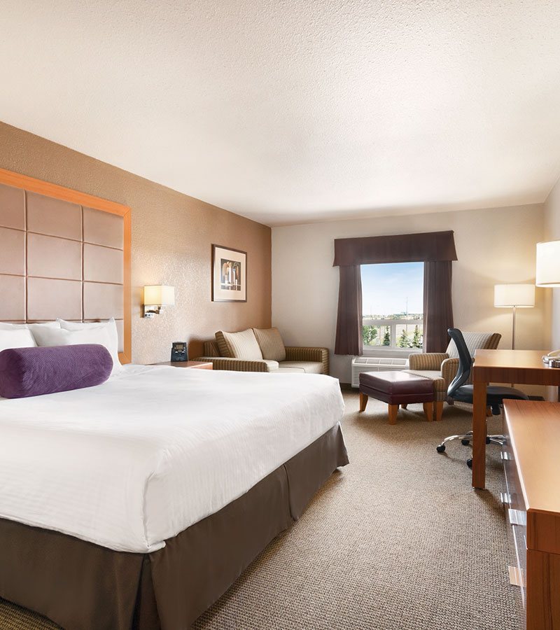 The Business Class suite at d3h Days Inn Calgary Airport features an impeccably made king bed along with a solid wood dresser, honey wood color writing table, tan armchair paired with a leather ottoman and a loveseat placed in a corner of the room.