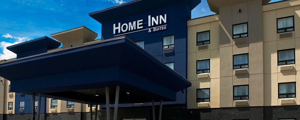 An upfront view of the flat roof navy blue portico that provides coverage over the entrance to the d3h Home Inn & Suites Saskatoon South.  The corporate name, in bold white typeset is displayed as signage high above the hotel entrance and portico.