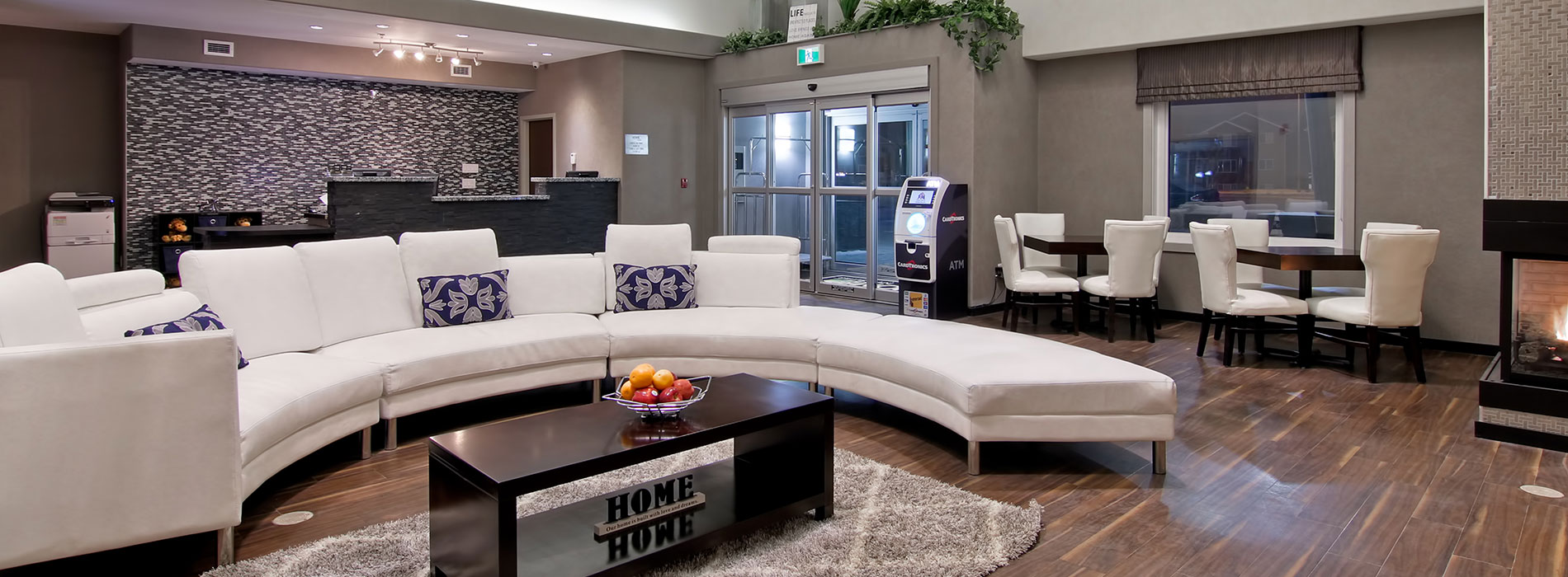 A large semi-circular white sectional sofa dominates the space in the lobby of d3h Home Inn & Suites Regina Airport.  Around the perimeter of the space is an ATM placed next to the glass door entrance and the slate gray stone check-in counter stands in stark contrast against a black, white and gray patterned accent wall.