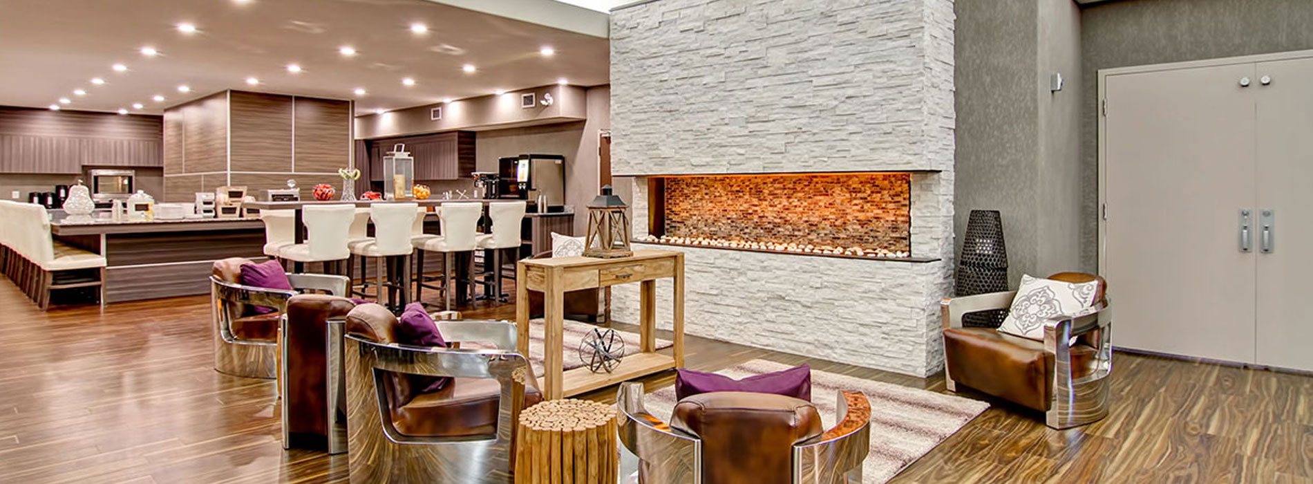 A towering birch white limestone electric fireplace is surrounded by leather club chairs, tree stump side tables, area rugs and a rustic console table bearing metal and wood décor items in the lounging space at d3h Home Inn & Suites Swift Current.  In the background, the wood grain laminate interior of the breakfast area can be seen among neat rows of white upholstered Parsons dining chairs.