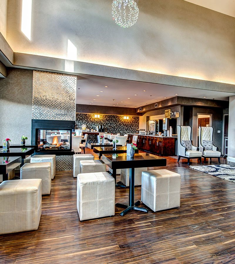 Square thick dark wood eating tables and seating options including white cube pouf ottomans and white Parsons dining chairs populate the hardwood floor HomeEssentials breakfast area at d3h Home Inn & Suites Swift Current.  The space is dominated by a glass enclosed fireplace with a silver shiny metal surround and a elongated mahogany buffet that serves as the breakfast counter.