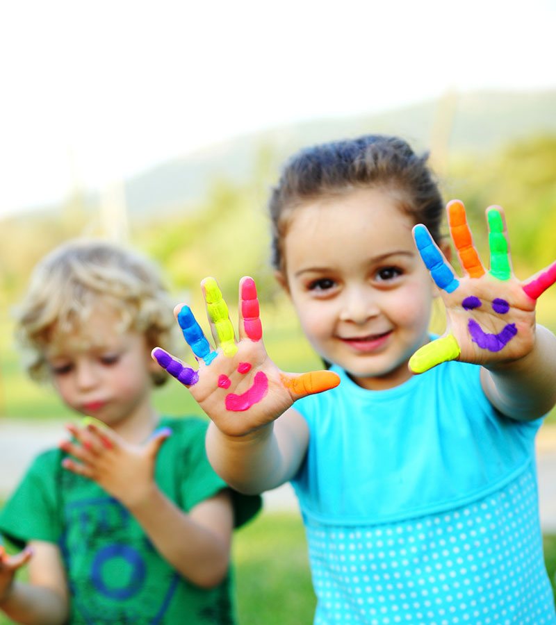 A young girl is eagerly and happily holding up her fingers painted in bright multiple colors with palms painted with a happy face, while in the background a younger blond male child stares contemplatively at his own painted hands.