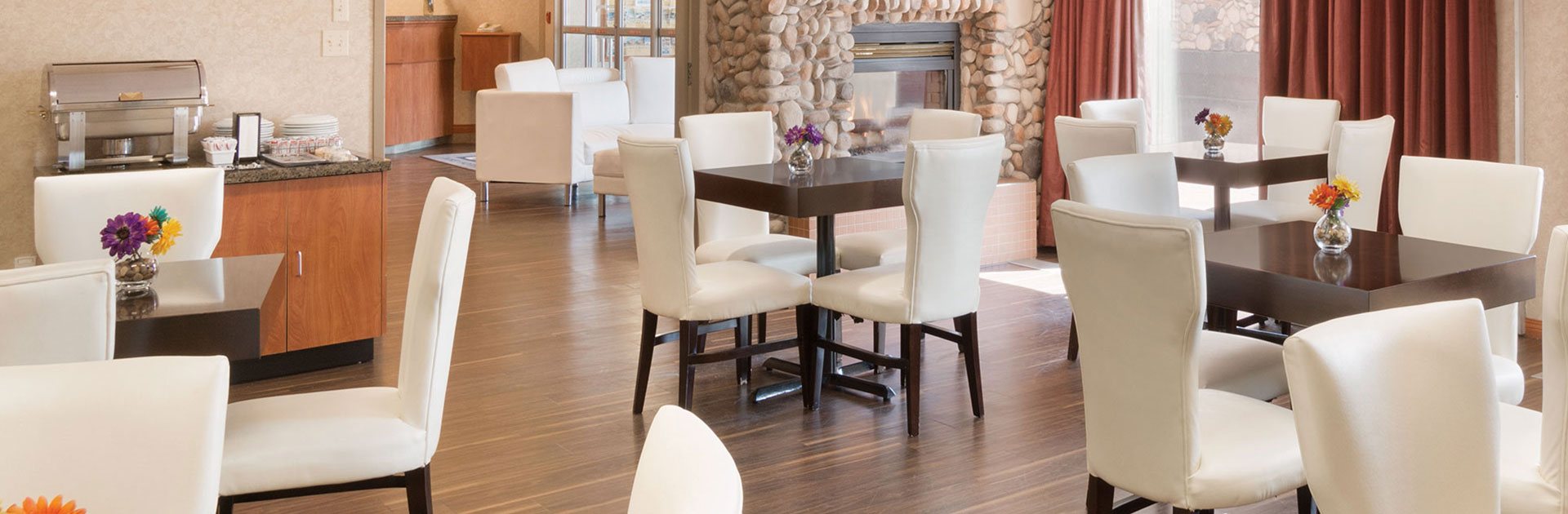 Black brown square eating tables with white cube shaped pouf ottoman seating are placed around a burning electric fireplace inset with black trim at the breakfast room at d3h Home Inn & Suites in Yorkton, Saskatchewan.