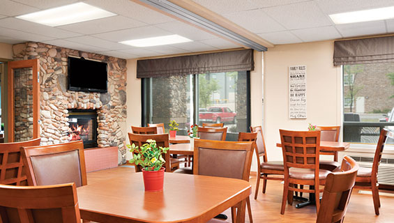 The dining room at d3h Days Inn Calgary Airport centres around a stone fireplace with a brick base and a flatscreen TV mounted above the hearth.  Square honey brown eating tables with matching wood and leather upholstered chairs are placed throughout the space, while windows and a sliding glass patio door allows natural light to flow in.