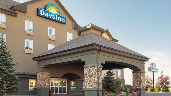 Two tall spruce trees are placed by a concrete and stone portico built over the glass door entry to d3h Days Inn Medicine Hat in Alberta.  At the top of the triangular roof of the hotel, the Days Inn corporate logo, presented in white lettering and yellow sunrise icon set against a blue background, is displayed over the entrance.