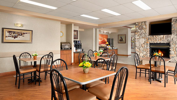 The eating and dining area of the Daybreak Café at d3h Days Inn Red Deer is kept warm by a burning stone fireplace, with a flatscreen TV sitting directly above the hearth.  Square bronze brown eating tables (with plastic centrepieces filled with flowers) and upholstered chairs are placed throughout the breakfast space.  In the corner of the room, a food warming appliance and a coffee kiosk sit on top of red wood stained counters.