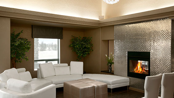 The cozy lounging space at d3h Days Inn Regina showcases a semi-circular white sectional couch placed by a window looking out into the wintry scenery.  The couch extends next to the glass enclosed fireplace encased by a silver metallic herringbone pattern surround.  Additional seating is provided by two high back white upholstered Parsons chairs and a set of four cube shaped tan ottomans.