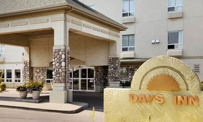 A legacy photograph of a d3h Days Inn property in 1999.  The glass front entrance is covered over by a square concrete portico with stone accent trim on the support posts.  Concrete planters bear plants and flowers and a yellow stone monument of a sunset icon of the corporate logo stands in the foreground.