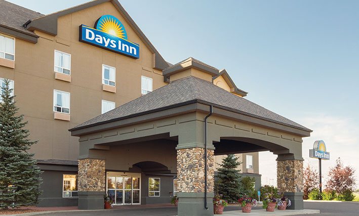 A large pavilion shaped concrete portico structure with stone accents is built over the entrance to the d3h Days Inn Medicine Hat in Alberta.  At the topmost point of the building, the corporate logo (bright yellow sunrise icon and brand name in bold white lettering against a blue background) overlooks the entrance area adorned with terracotta planters of florals and two tall Douglas fir trees planted alongside the portico.