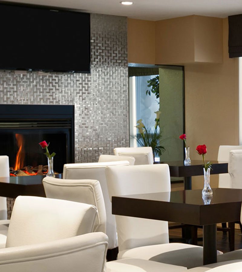 A view of the glass enclosed electric fireplace with a silver metal geometric pattern wall surround and mounted flatscreen TV in the dining area at d3h Days Inn Regina East in Saskatchewan.   White upholstered dining chairs are paired up with dark square eating tables each topped with a single rose in a vase.