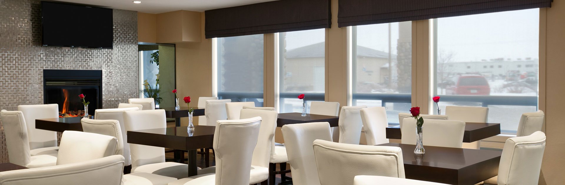 Four large windows let natural light into the dining space of d3h Days Inn Regina populated with square penny brown wood tables, topped with a single red rose in a glass bud vase, amidst a sea of white upholstered dining chairs.  The eating space is dominated by the glass enclosed fireplace with a shiny silver metallic surround and a flatscreen TV mounted above.