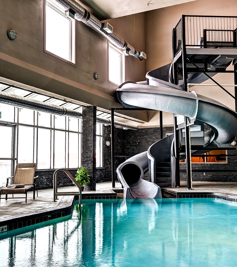 A wicker chaise lounge is positioned next to the twisting waterslide and indoor pool at d3h Home Inn & Suites in Swift Current, Saskatchewan.  A wall of windows and two square windows positioned high above the pool area provide illumination into the dark gray stone brick interior.