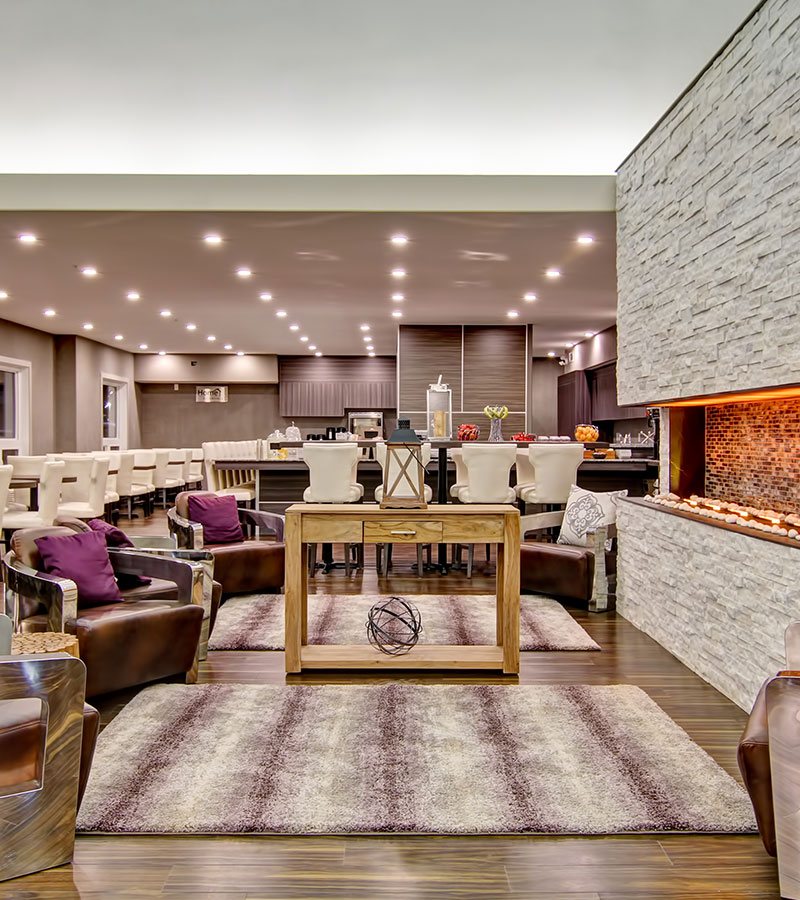 Leather club chairs adored with purple accent pillows encircle a white limestone electric fireplace at the lounging space at d3h Home Inn & Suites Saskatoon South.  Eating tables and dining benches, all surrounded by white upholstered Parsons dining chairs, fill the eating area, with a view of the woodgrain laminate panels of the breakfast bar in the background.
