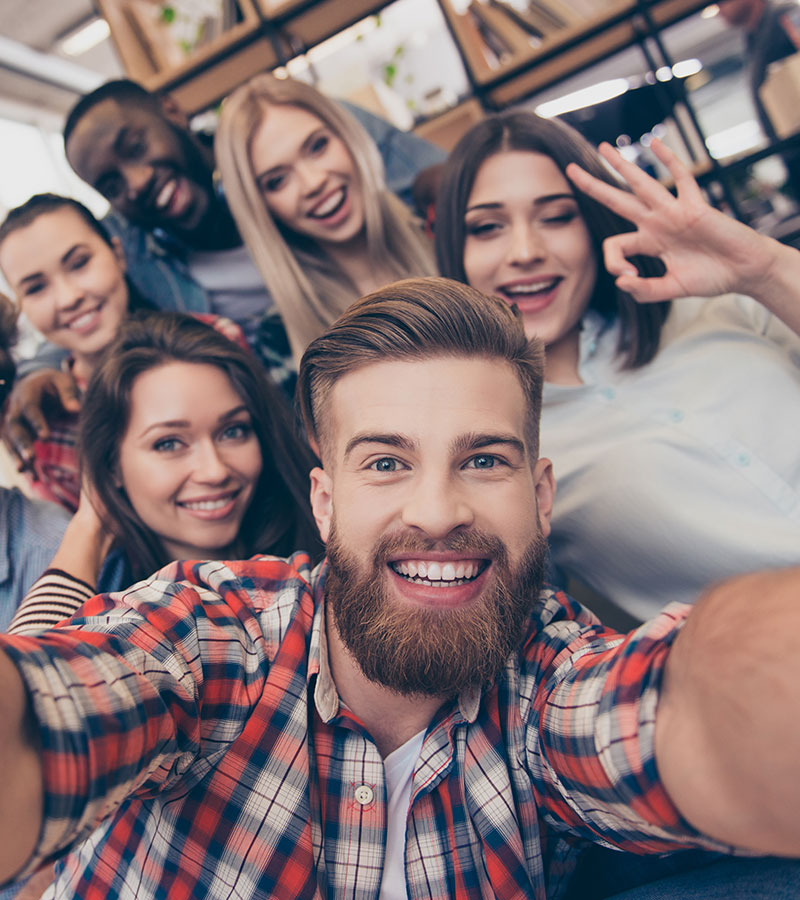 A happy group of young men and women pose for a group selfie.  The brown haired bearded young male of the group holds up the camera.