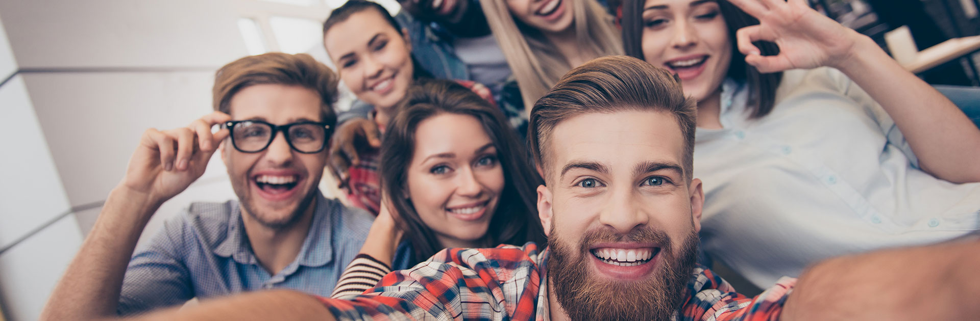 A group of young people smiling and posing for a selfie with a young bearded man in a multi-colored plaid shirt in the forefront holding up a camera phone.