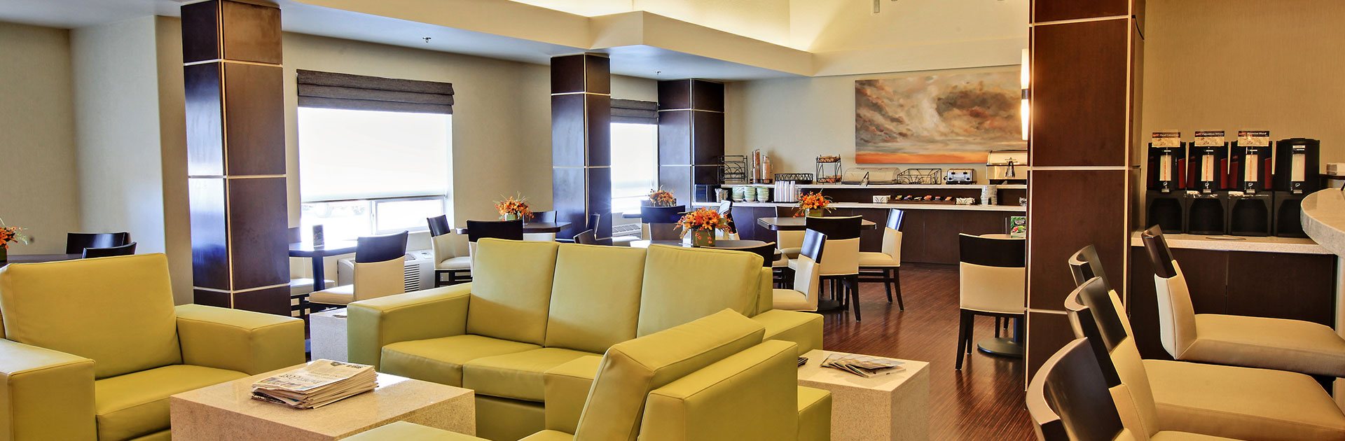 A chartreuse green sofa and armchair set, paired with a solid white cube shaped ottoman, defines the lounge space at d3h HomeSuites Regina East.  Adjacent to the lounging area, is the breakfast space defined by eating tables and white upholstered dining chairs placed alongside dark wood paneled columns.