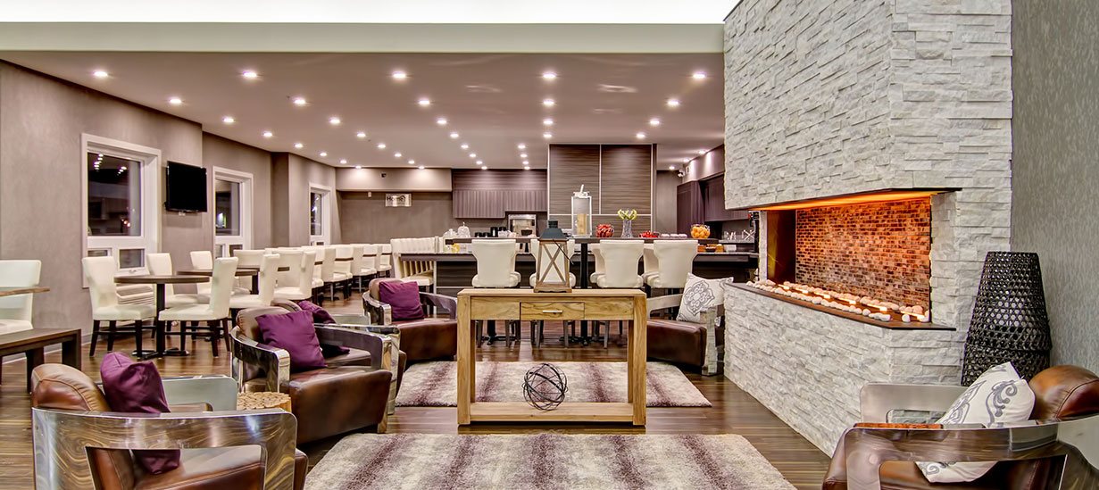 A panoramic view of the dining and lounge areas of d3h Home Inn & Suites Saskatoon South featuring a white stacked limestone electric fireplace encircled by leather club chairs, with rows of eating tables and clusters of white upholstered dining chairs placed throughout the dining space.  The eat-in breakfast bar, surrounded by additional white dining chairs, is stocked with fruit and beverages.
