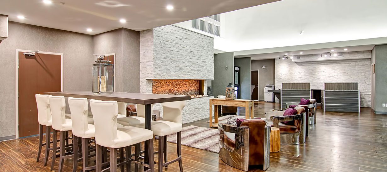 A long dining bench surrounded by white upholstered Parsons dining chairs overlooks the view of wood grain laminate check-in counters and the lounging space with leather club chairs encircling a majestic white stacked limestone electric fireplace at d3h Home Inn & Suites Saskatoon South.  