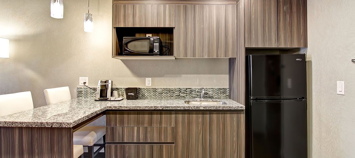 The kitchenette in the Suite Dream Room at d3h Home Inn & Suites Saskatoon South is equipped with mocha brown wood grained laminate panel cabinets, a marbled granite top eat-in breakfast bar with two white upholstered dining chairs, and includes amenities such as coffee maker, microwave and a black refrigerator.
