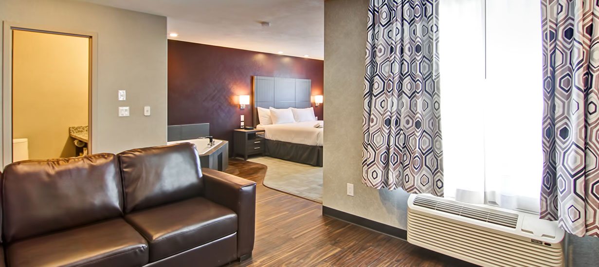 The Jacuzzi Room at d3h Home Inn & Suites Saskatoon South showcases a hot tub placed next to a king sized bed and an accompanying chocolate brown wood beside table.  A dark brown leather loveseat is placed next to an air conditioning unit and a window draped with geometric print curtains.
